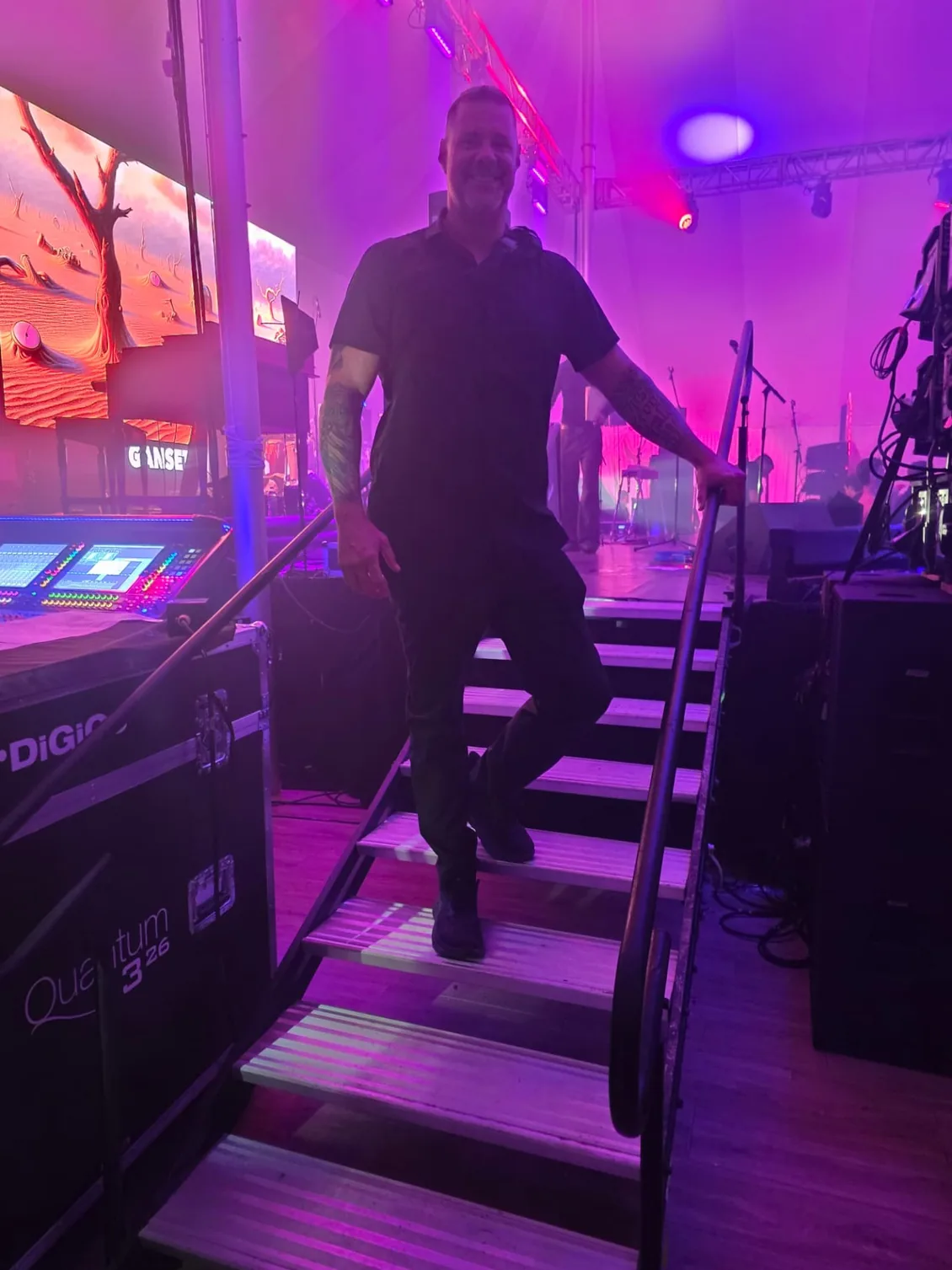 Protection agent on the stage stairs beside audio consoles at a tented live production under purple lighting