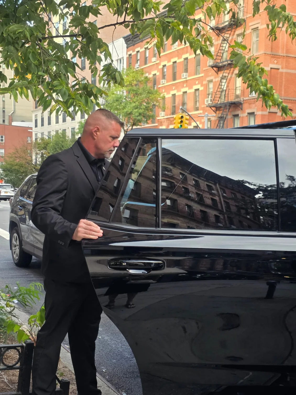Shadow Securities protection agent holding the rear door of a black SUV at a Manhattan curb on a tree-lined street