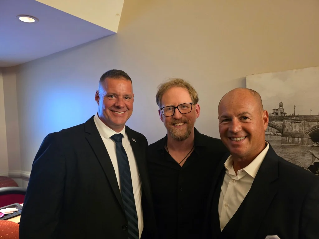 James Gonzales, owner of Shadow Securities, in a dark suit standing with two men at a private indoor engagement.